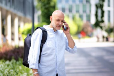 50 'li yaşlarda şehir caddesinde yürüyen kendine güvenen bir adam. Kel ve sakallı erkek, gömlek ve siyah pantolon giyiyor. Omzunda siyah bir sırt çantası ve elinde bir akıllı telefon vardı. Modern ofis binaları ve arka planda yeşil konuşmalar.
