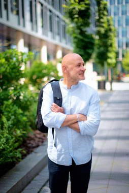 50 'li yaşlarda şehir caddesinde yürüyen kendine güvenen bir adam. Kel ve sakallı erkek, gömlek ve siyah pantolon giyiyor. Omzunda siyah bir sırt çantası vardı. Modern ofis binaları ve arka planda yeşil konuşmalar.