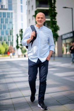 50 'li yaşlarda şehir caddesinde yürüyen kendine güvenen bir adam. Kel ve sakallı erkek, gömlek ve siyah pantolon giyiyor. Omzunda siyah bir sırt çantası vardı. Modern ofis binaları ve arka planda yeşil konuşmalar.