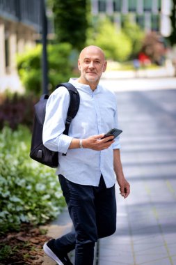 50 'li yaşlarda şehir caddesinde yürüyen kendine güvenen bir adam. Kel ve sakallı erkek, gömlek ve siyah pantolon giyiyor. Omzunda siyah bir sırt çantası ve elinde bir akıllı telefon vardı. Modern ofis binaları ve arka planda yeşil konuşmalar.
