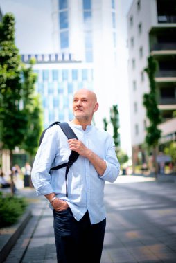 50 'li yaşlarda şehir caddesinde yürüyen kendine güvenen bir adam. Kel ve sakallı erkek, gömlek ve siyah pantolon giyiyor. Omzunda siyah bir sırt çantası vardı. Modern ofis binaları ve arka planda yeşil konuşmalar.