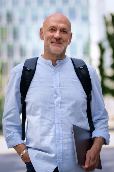 50 'li yaşlarda şehir caddesinde yürüyen kendine güvenen bir adam. Kel ve sakallı erkek, gömlek ve siyah pantolon giyiyor. Omzunda siyah bir sırt çantası ve elinde bir dizüstü bilgisayar vardı. Modern ofis binaları ve arka planda yeşil konuşmalar.