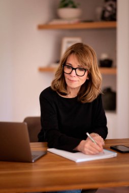 Kendine güveni tam orta yaşlı bir kadın modern bir ofis masasında rahatça oturuyor. Siyah kazak giyen ve iş için laptop kullanan çekici bir kadın.. 