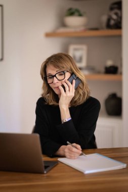 Kendine güveni tam orta yaşlı bir kadın modern bir ofiste rahat bir şekilde masasında oturuyor ve dizüstü bilgisayarını kullanıyor. Beyaz gömlek giyen ve elinde akıllı telefon tutan çekici bir kadın.. 