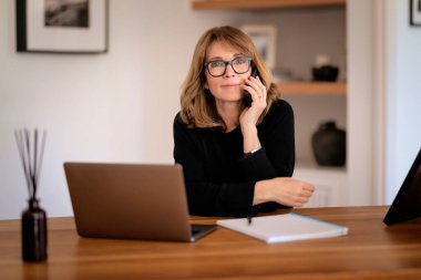 Kendine güveni tam orta yaşlı bir kadın modern bir ofiste rahat bir şekilde masasında oturuyor ve dizüstü bilgisayarını kullanıyor. Beyaz gömlek giyen ve elinde akıllı telefon tutan çekici bir kadın.. 