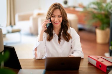 Kendine güveni tam orta yaşlı bir kadın modern bir ofis masasında rahatça oturuyor. Beyaz gömlek giyen ve elinde akıllı telefon tutan çekici bir kadın.. 