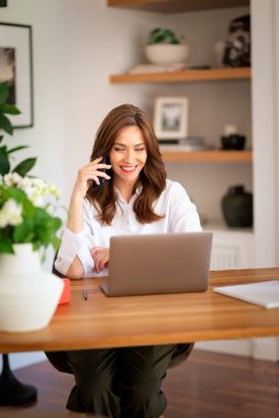 Kendine güveni tam orta yaşlı bir kadın modern bir ofis masasında rahatça oturuyor. Beyaz gömlek giyen ve elinde akıllı telefon tutan çekici bir kadın.. 