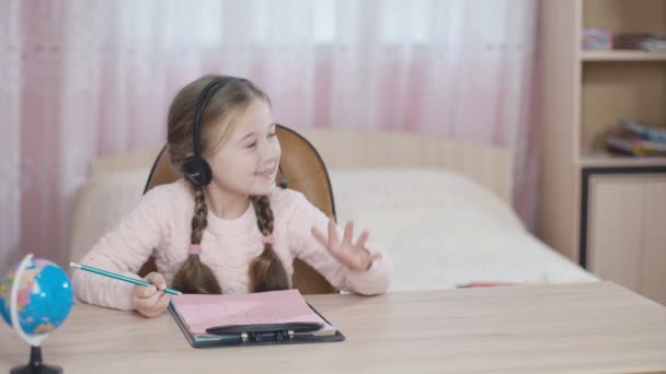 Маленькая девочка в наушниках сидит за столом — стоковое видео