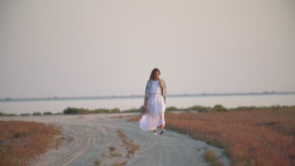 Chica con flores en sus manos va en un camino arenoso — Vídeos de Stock