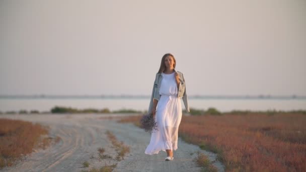 Девушка с цветами в руках идет по песчаной дороге — стоковое видео