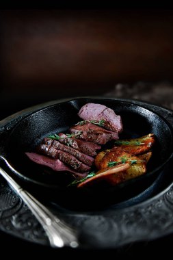 Pan-fried wild venison fillet with fried potato slices and thyme herbs. Shot against a dark, rustic background with generous accommodation for copy space. clipart