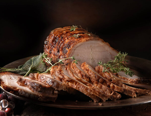 A perfectly oven roasted prime pork joint carved and ready to serve with thyme and sage herbs. Shot against a dark rustic background with generous accommodation for copy space.