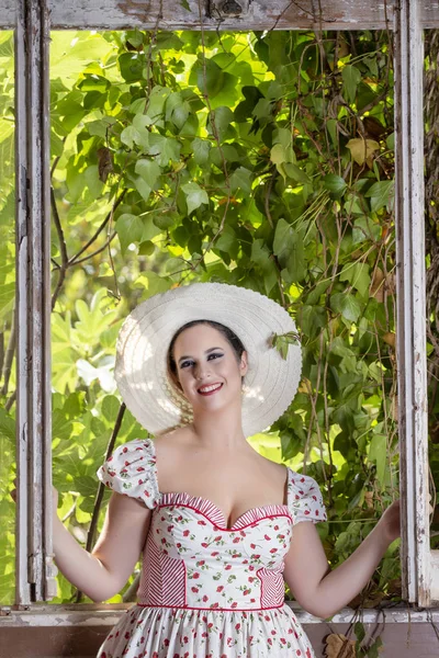 看到一个乡村的年轻淑女妇女与白色的礼服和红色的花朵和帽子在一个废弃的宫殿旁边的窗口 — 图库照片