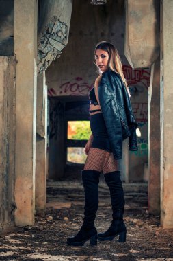 Terk edilmiş bir fabrika binasında deri ceket, siyah bluz ve kısa kot pantolon giyen genç bir kız..