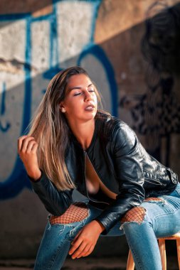 Deri ceketli, mavi kotlu, terk edilmiş bir fabrikada şehvetli pozlar veren bir model..