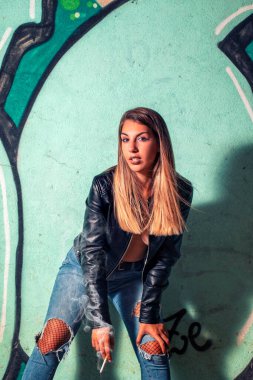 Deri ceketli ve kot pantolonlu bir manken terk edilmiş bir fabrikada şehvetli pozlar veriyor, puro içiyor..