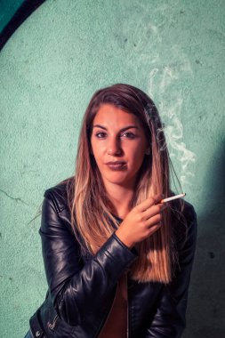 Deri ceketli ve kot pantolonlu bir manken terk edilmiş bir fabrikada şehvetli pozlar veriyor, puro içiyor..