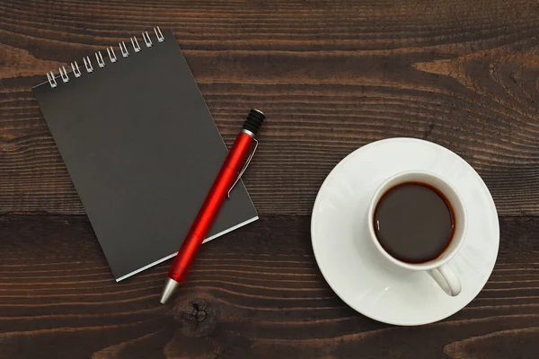 Cup of coffee, pen and black notebook on a wooden background. View from above.