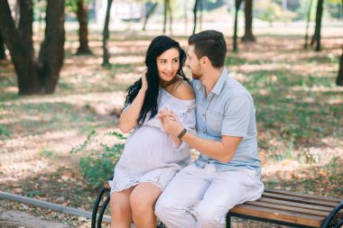 Genç bir adam ve kız arkadaşı hamile yeşillik ve ağaçlar arka planı bir bankta oturuyor. Bir çocuk beklentisiyle. İlk gebelik seviyorum çocuklar.