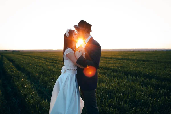 美丽的新娘和时尚的新郎亲吻在田野里 新郎在帽子 夕阳的光芒之间的夫妇 — 图库照片