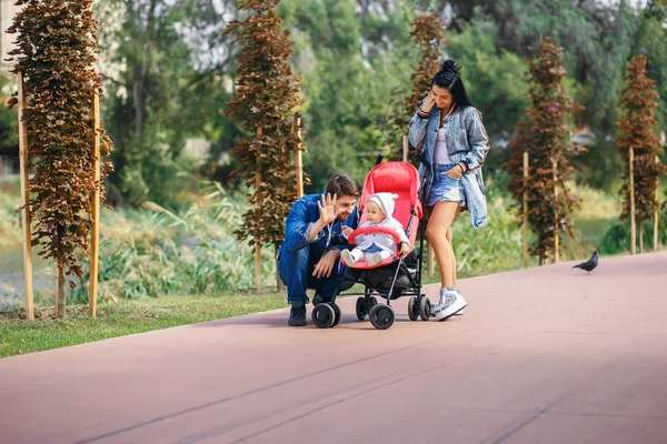 Mamá y papá están caminando en el parque con su hija en un cochecito, un hermoso día soleado, una hermosa familia — Foto de Stock