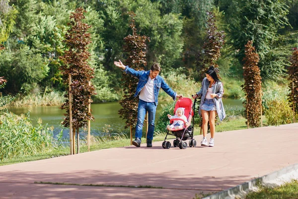 Mama und Papa spazieren mit ihrer Tochter im Kinderwagen durch den Park, ein schöner sonniger Tag, eine schöne Familie — Stockfoto