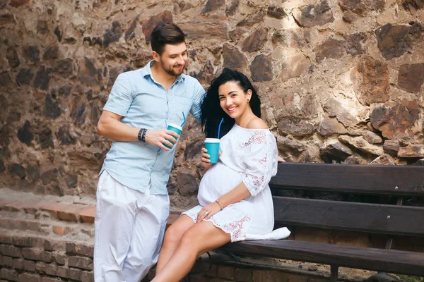 Hermosa pareja está sentada en un lugar romántico, sonriendo y besándose, bebiendo té caliente. Embarazada esposa y su marido, pared de piedra y banco —  Fotos de Stock