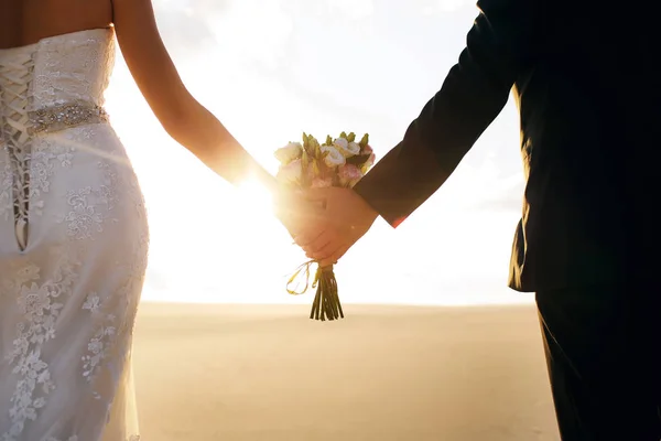 A menyasszony és a vőlegény kéz a kézben, a kezében egy szép csokor, a pár állt a sivatagban, a naplemente — Stock Fotó