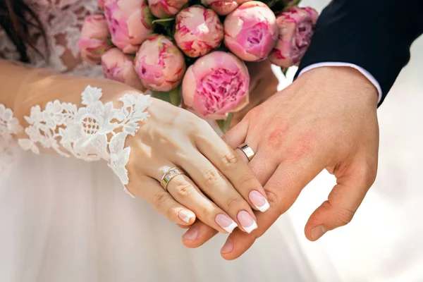 Handen van de pasgetrouwden met ringen op de vingers, naast een boeket met roze pioenrozen, de bruid en bruidegom handen te houden — Stockfoto