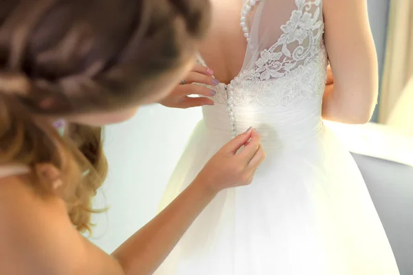 Dama de honra ajudando noiva esbelta amarrando seu vestido branco do casamento, abotoando no padrão de renda delicada com saia macia na cintura. Manhã nupcial preparação detalhes recém-casados. Momentos do dia do casamento, desgaste — Fotografia de Stock