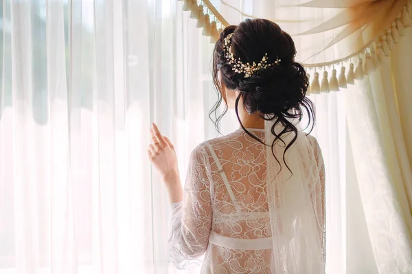 Visão traseira de uma noiva morena elegante vestida com um vestido branco, a menina olha pela janela. Conceito de casamento — Fotografia de Stock