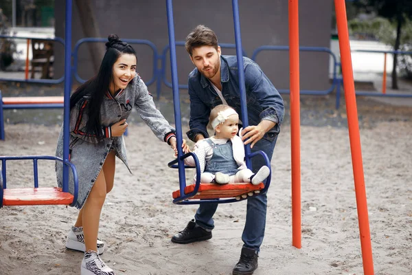 Schöne junge Eltern, spazieren gehen mit einem Kind, schaukeln es auf einer Schaukel, haben Spaß und genießen einander, glückliche Familie für einen Spaziergang — Stockfoto