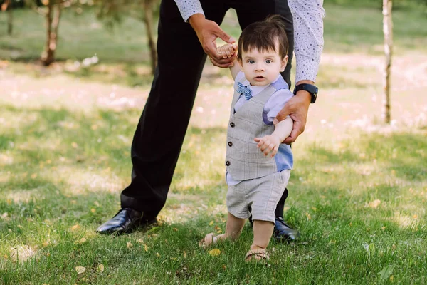 father teaches his little son to walk, family walks in the park, dad holds the child's hands and helps to walk, evening walk, father-son relationship, trust in the family, beautiful baby