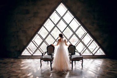 Gelin, iki çizgili koltuğun yanında gelinlik içinde, arka planda üçgen bir pencere, şık bir manzara, klasik bir iç mekan, inanılmaz bir pencere, güzel bir tasarım.