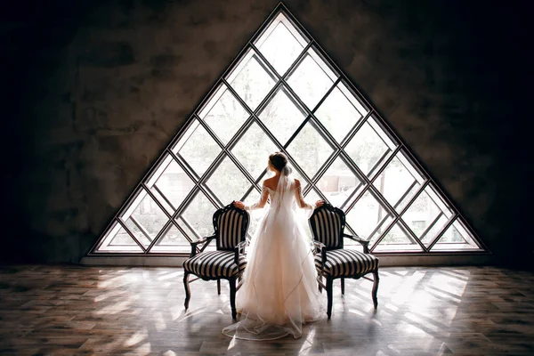 Gelin, iki çizgili koltuğun yanında gelinlik içinde, arka planda üçgen bir pencere, şık bir manzara, klasik bir iç mekan, inanılmaz bir pencere, güzel bir tasarım.
