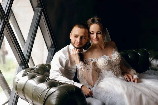 新娘和新郎坐在窗边的沙发上拥抱 结婚纪念日 漂亮的白色连衣裙 爱情的气氛 婚礼仪式 时髦的丈夫和乌黑的妻子 — 图库照片
