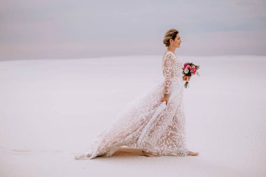 Güzel gelin çölde elinde bir buket, uzun beyaz gelinlik, mesaj için boş alan, mutlu ve gülümseyen sarışın, düğün günü konsepti, zayıf kız.