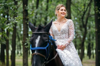 Güzel sarışın gelin düğün törenine siyah bir atla gidiyor, kız beyaz bir gelinlik giyiyor, güzel yeşil orman resmi bir parti için, mutlu kadın damada bakıyor.