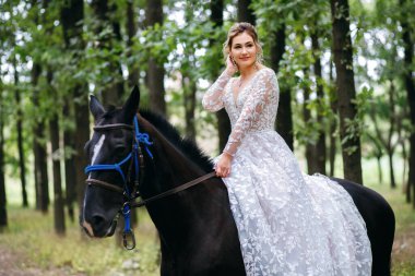 Güzel sarışın gelin düğün törenine siyah bir atla gider, kız beyaz bir gelinlik giyer, mesaj için boş alan, resmi bir parti için güzel yeşil orman, mutlu bir kadın.