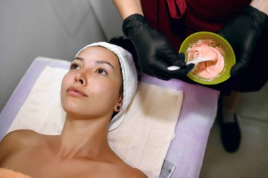 Esmer bir kız yüz ve boyun derisine maske takmak için prosedüre hazırlanıyor. Güzellik uzmanı malzemeleri ve kremleri karıştırıyor, cildine özen gösteriyor ve genç kalmasını sağlıyor.