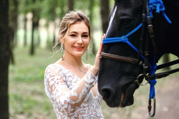 Beautiful bride in white wedding dress stroking a horse, portrait of a blonde with a black stallion, preparing to meet the groom on the wedding day, blonde with curls, place for text, wedding concept