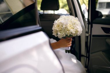 Beyaz elbiseli gelin, düğün arabasından iniyor ve elinde bir buket beyaz çiçek tutuyor, düğün günü, hiçbir yüz görünmüyor, mesaj için boş yer var.