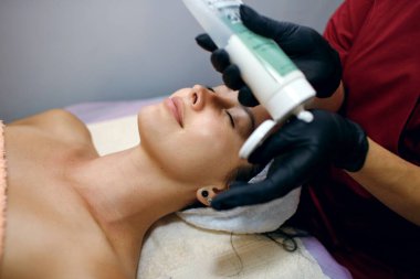 Kaplıca salonundaki esmer bir kadının yakın çekimi profesyonel bir kozmitolog müşterinin yüzüne krem sürer. Yüz bakımı ve vücut sağlığı kavramı