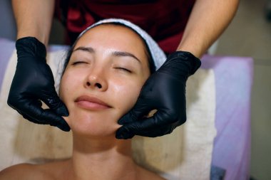 Güzellik merkezinde yüz masajı yaptıran genç bir kadının yakın çekimi. Cilt ve vücut bakımı için modern kozmetoloji. Siyah eldivenli profesyonel masör yüz masajı yapar.