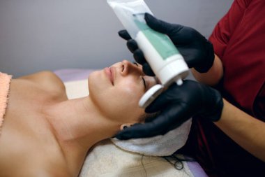 Kaplıca salonundaki esmer bir kadının yakın çekimi profesyonel bir kozmitolog müşterinin yüzüne krem sürer. Yüz bakımı ve vücut sağlığı kavramı