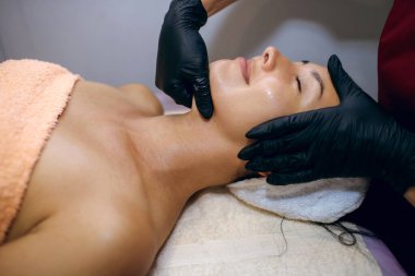 Güzel bir esmer kaplıcada tedavi görüyor, siyah mühürlü bir güzellik uzmanı yüzüne nemlendirici sürüyor. Yüz bakımı konsepti, profesyonel kaldırma, metin için boş alan