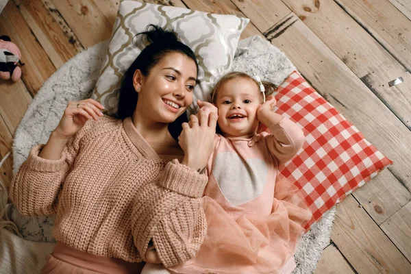 A beautiful brunette woman lies on a warm wooden floor and plays with her daughter. Family having fun together, top view of happy people, motherhood relationship concept