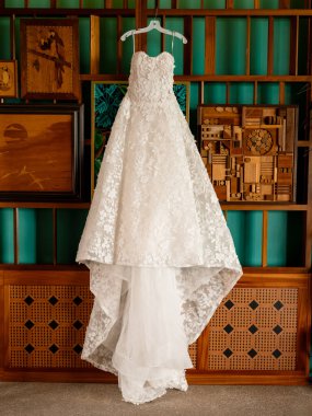 Uzun etekli zarif dantelli gelinlik. Beyaz gelinlik, evdeki bir odada yeşil bir duvarda asılı duruyor. Klasik gelinlik özel bir tören için hazırlandı.