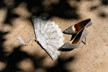 Bir çift parlak gümüş topuklu ayakkabı ve taştan bir yüzeye yerleştirilmiş inci saplı zarif bir dantel el yelpazesi. Düğün ya da lüks moda konsepti için mükemmel. Sanat fotoğrafları kadın aksesuarları.