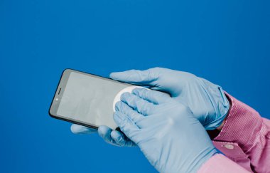 Tıbbi eldivenli bir adam telefonunu pamuk pedlerle siler. Mavi arka plan. Antibakteriyel etki, dezenfekte, dezenfekte